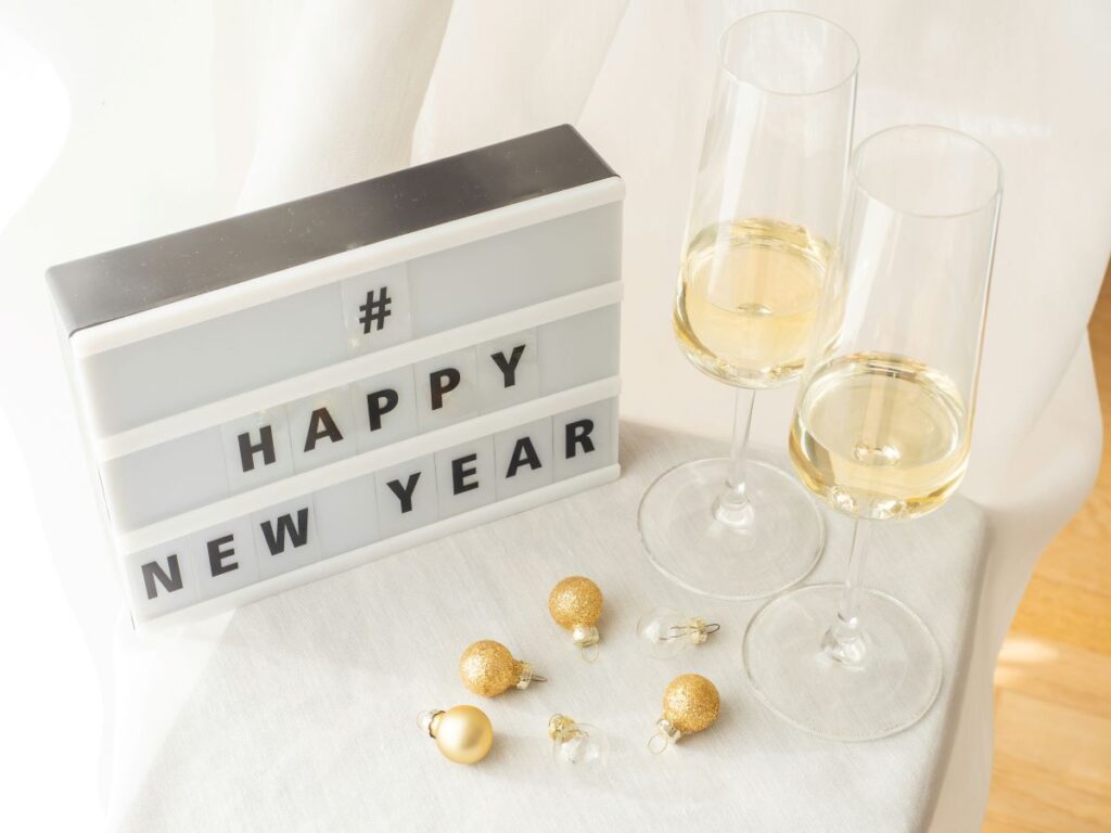 Two glasses of champagne, some gold, sparkly ornaments and a sign-box that says #Happy New Year